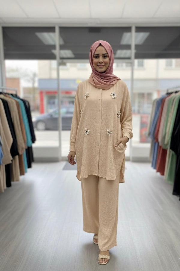 Une femme portant une tunique ample à manches longues en tissu gaufré de couleur beige clair et un pantalon large assorti. La tunique est boutonnée sur le devant et ornée de broderies décoratives en forme de coquillages ou de fleurs. La femme porte également un hijab rose clair et des sandales de couleur claire. L'arrière-plan est un magasin de vêtements.