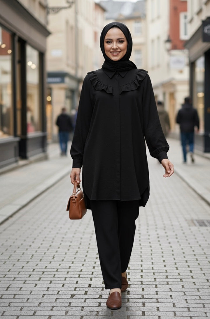 Femme souriante portant un ensemble noir élégant avec une tunique à volants et un pantalon assorti, marchant dans une rue pavée.