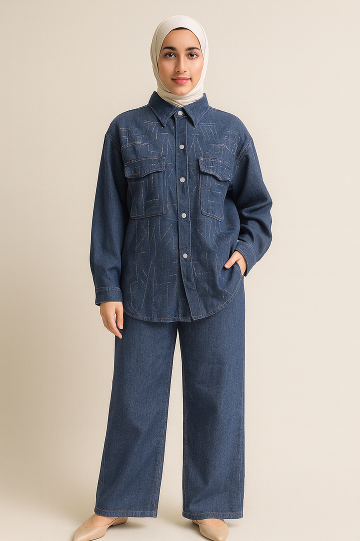 Femme voilée portant un hijab beige clair et un ensemble en denim bleu 2 pièces composé d’un pantalon large et d’une chemise boutonnée avec surpiqûres décoratives, posant devant un fond beige clair. Disponible en tailles S, M, L et XL.