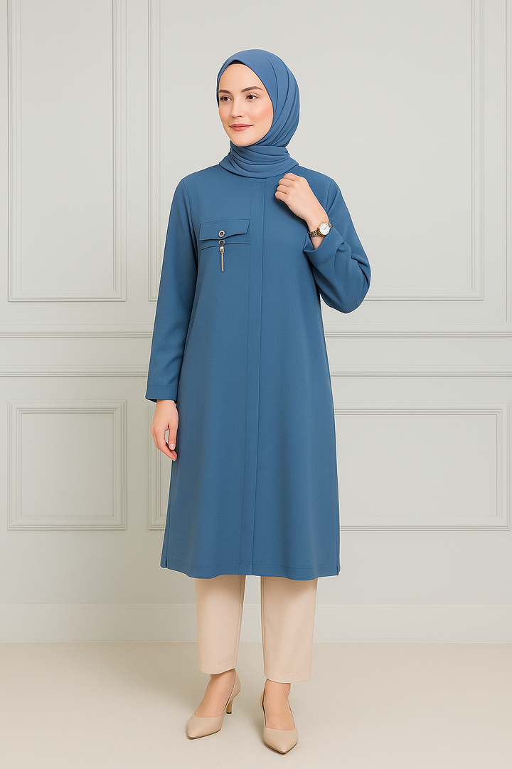 Femme portant une tunique longue bleu pétrole avec poche décorative et zip, manches longues, associée à un hijab bleu assorti et un pantalon beige clair.