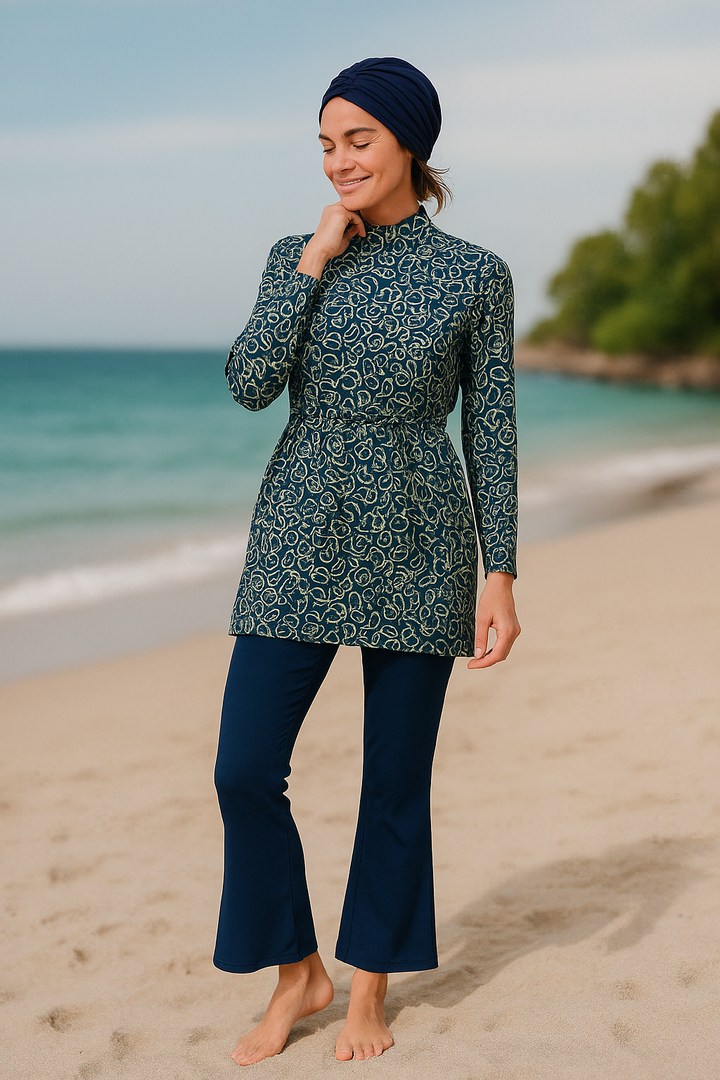 Femme élégante en burkini imprimé bleu et vert, avec tunique cintrée à motifs et pantalon évasé, marchant sur une plage paisible.